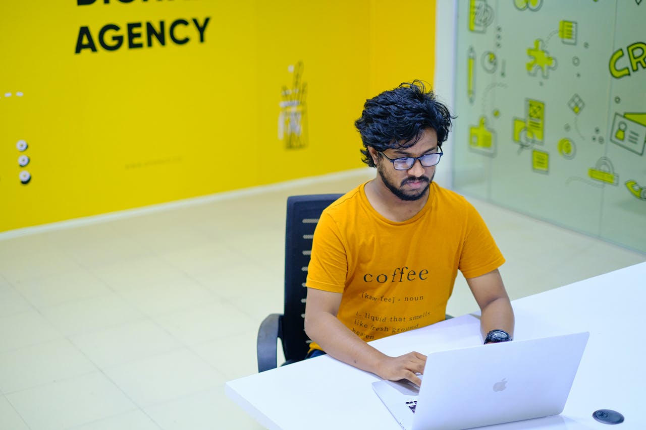hero-img-01 Young man wearing eyeglasses working on laptop in vibrant digital agency office.